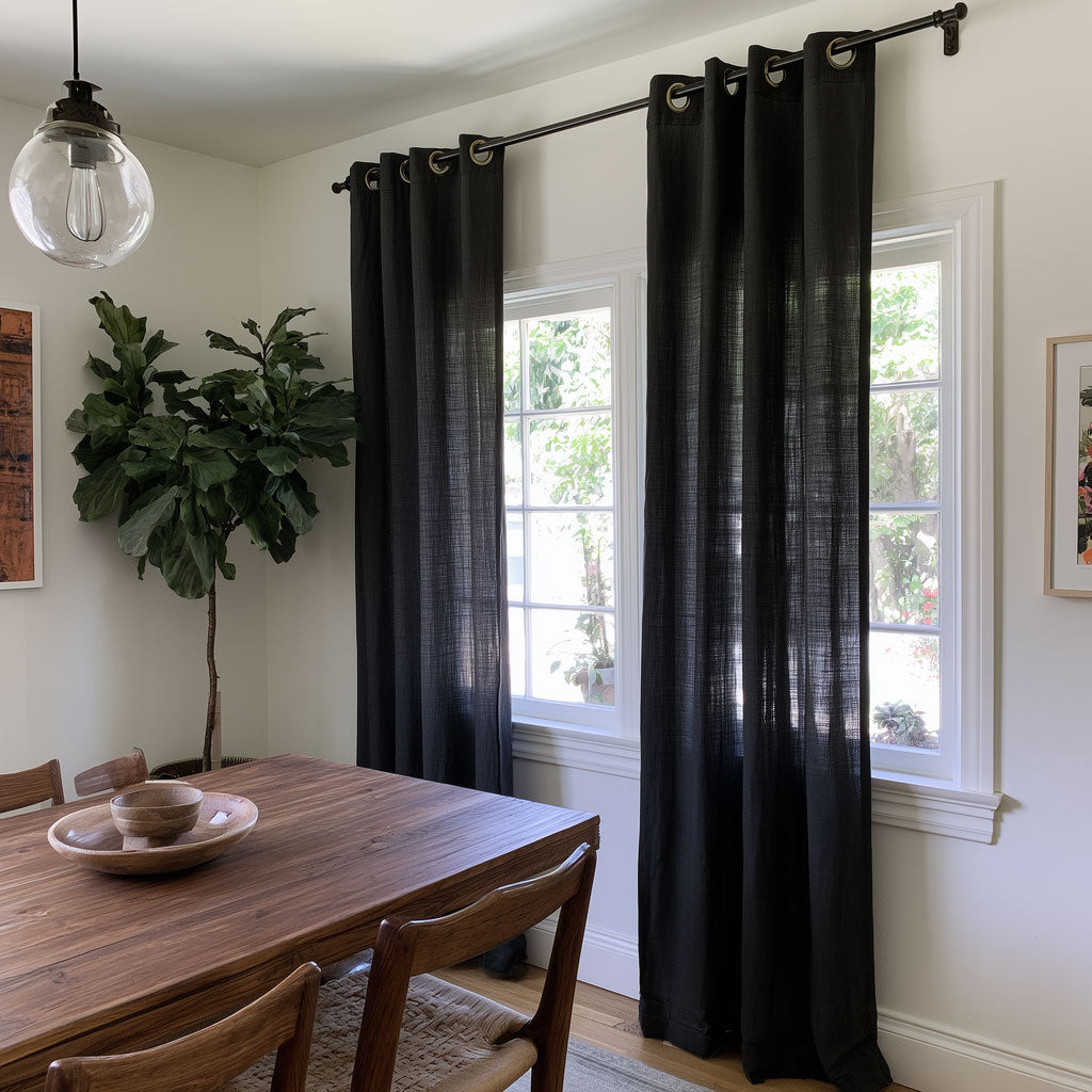 Floor-length black eyelet curtains hang beside two dining room windows, with a wooden table, woven-seat chairs, a tall leafy plant, and a glass pendant light.