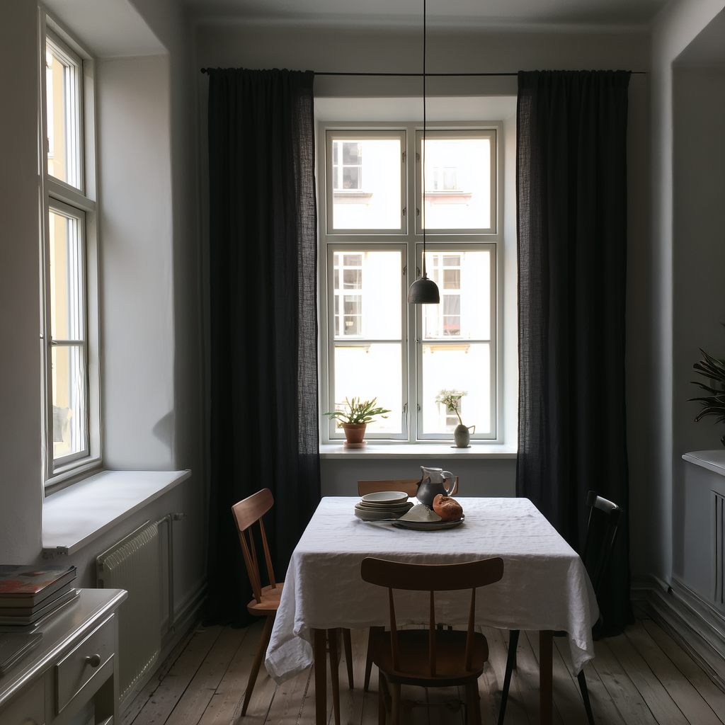Floor-length black curtains frame a tall window behind a small dining table with a white tablecloth, wooden chairs, stacked plates, a pitcher, and a black pendant light