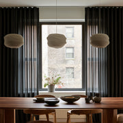 Floor-length charcoal gray curtains frame a large dining room window behind a wooden table with ceramic dishes, wooden chairs, and three cream pendant lights