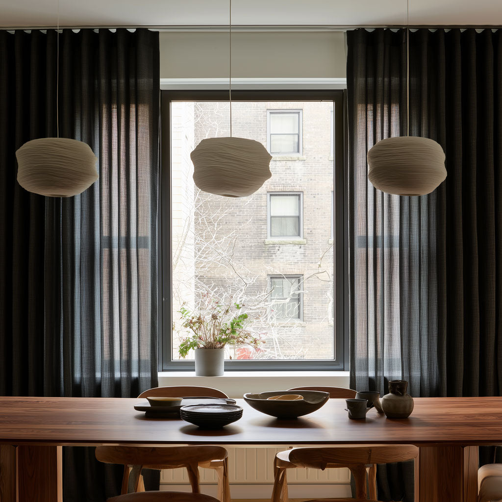 Floor-length charcoal gray curtains frame a large dining room window behind a wooden table with ceramic dishes, wooden chairs, and three cream pendant lights