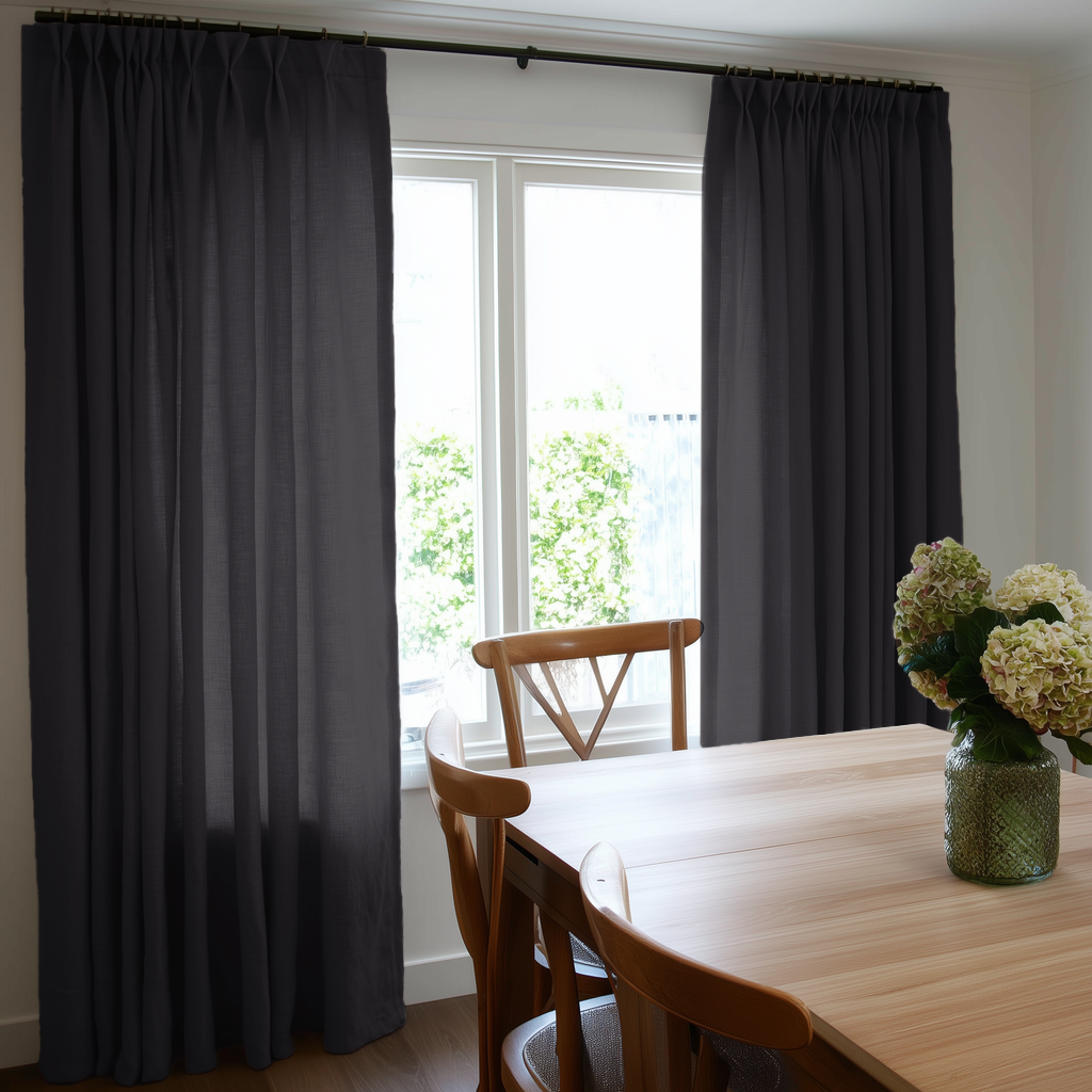 Floor-length charcoal gray pinch-pleat curtains frame a bright window beside a wooden dining table and chairs, with pale hydrangeas in a green glass vase