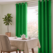 A fresh dining space with vibrant green curtains, a table covered in a neutral linen cloth, upholstered beige chairs, a simple tea setting, and a potted indoor tree