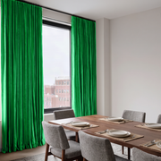 A modern urban dining room with vivid green curtains, a wooden table, textured gray chairs, minimalist place settings, and a city view through the large window