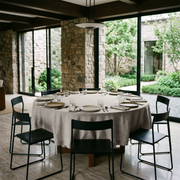 Natural Linen 108 Inch Round Tablecloth in a Modern Stone Dining Room with Floor-To-Ceiling Windows and Black Metal Chairs