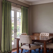 A sophisticated dining room with soft green curtains, French doors, a dark wooden table, upholstered chairs, neutral walls, and an elegant blue area rug
