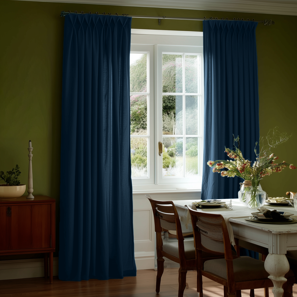 Floor-length teal blue pinch-pleat curtains frame a white window against olive green walls, beside a white dining table with wooden chairs, place settings, and a vase of flowers