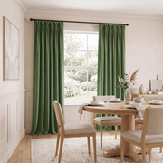 A bright, welcoming dining room with rich olive-green curtains, a round light-wood table, neutral upholstered chairs, simple tableware, and a delicate dried-floral centerpiece