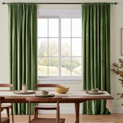 A bright minimalist dining room with floor-length olive-green curtains, a warm wooden table, simple wooden chairs, neutral ceramic tableware, and a soft garden view