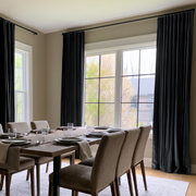 A bright contemporary dining room with large corner windows, elegant black curtains, a dark wooden table, upholstered taupe chairs, and neatly arranged tableware