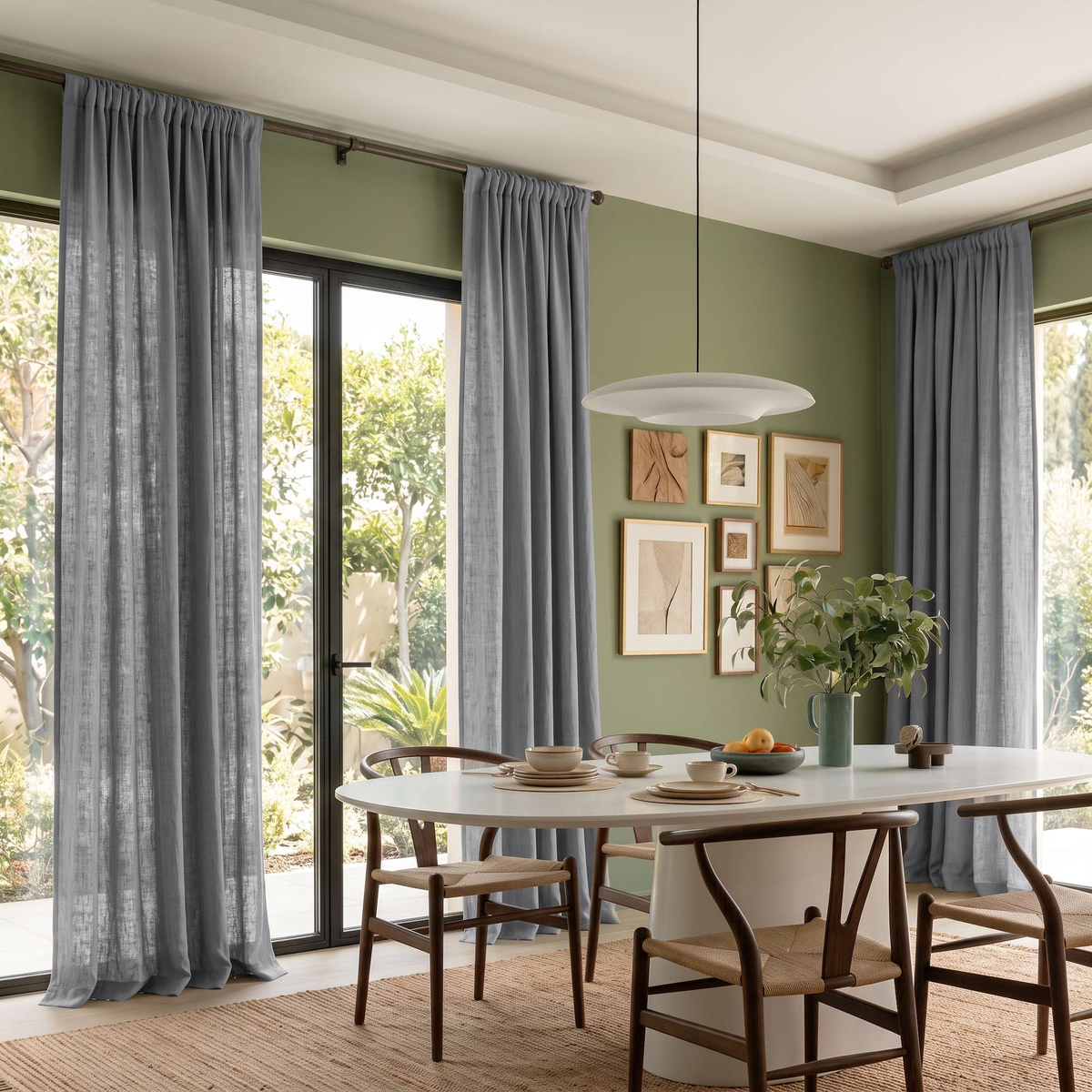 Dining room with sage green walls, floor-length gray curtains, a white oval table, wooden woven-seat chairs, and a pendant light, with greenery visible through large windows.