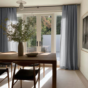 Floor-length gray curtains frame glass patio doors in a sunlit dining room with a wooden table, chairs, and a tall vase of leafy branches